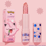 ColourPop pH Lip Balm Sing stands uncapped, revealing a clear glittery bullet beside its pink Pokémon x ColourPop box and matching cap with blue starbursts and Jigglypuff art, set against a pastel pink background.