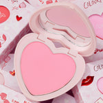 Sweetheart heart-shaped compact opens to reveal a soft baby pink pan and curved mirror, ColourPop Pressed Powder Blush, resting on Valentine-themed packaging with hearts, roses, and lip prints.